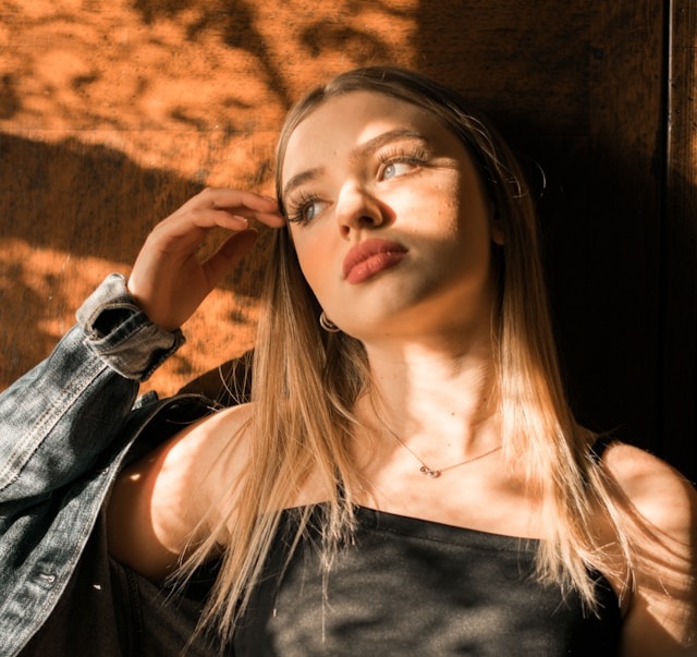A young woman with long blonde hair poses against a wooden wall, illuminated by warm, dappled sunlight while wearing a denim jacket and black top.