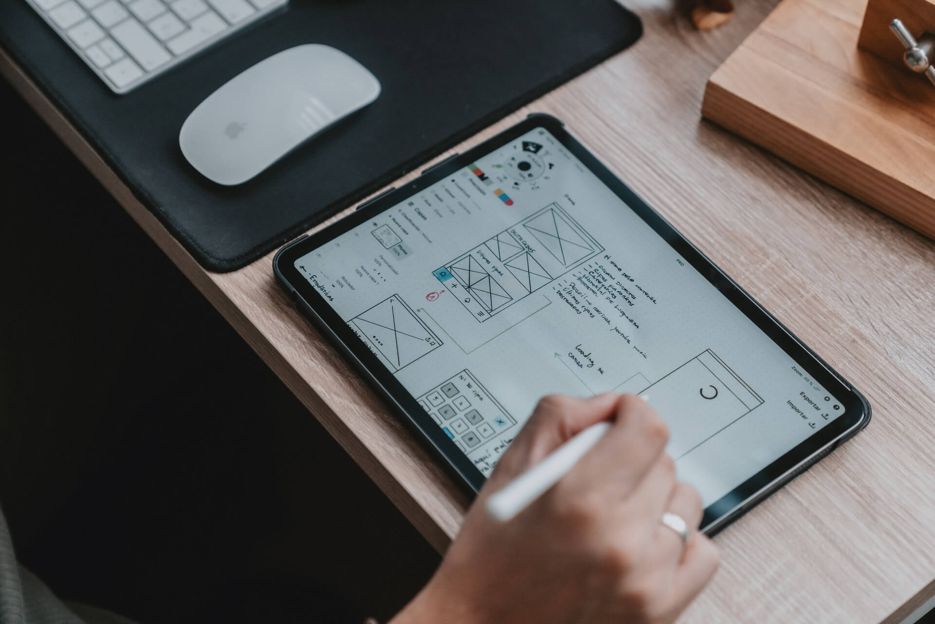 A person uses a stylus on a tablet to draw a website wireframe layout, with a computer mouse and keyboard visible on a wooden desk workspace nearby.