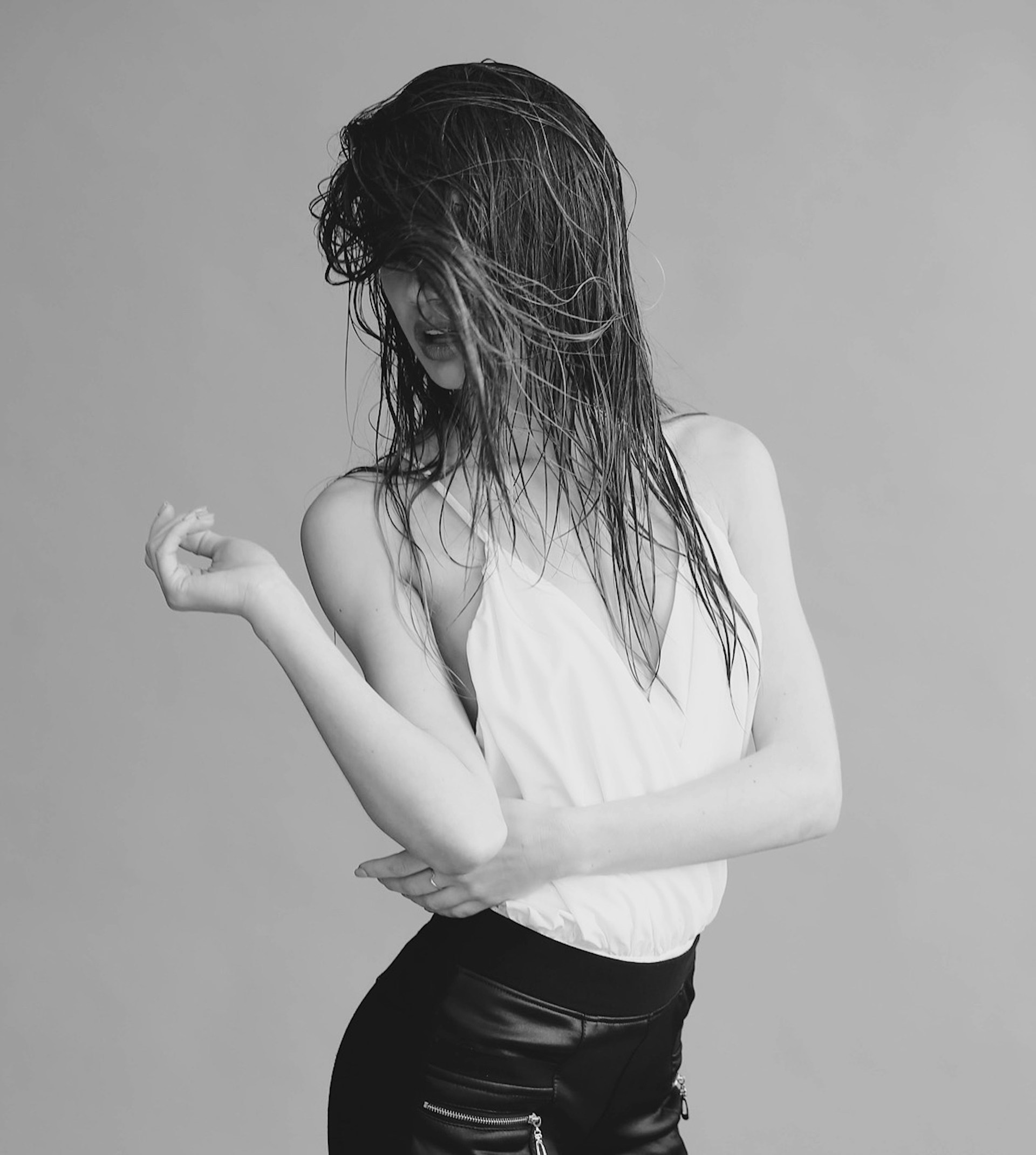 A black and white portrait of a woman with wet hair covering her face, wearing a white camisole and dark pants, posing against a plain background.