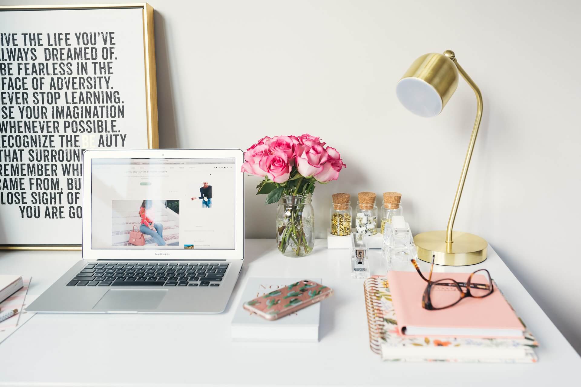 A clean white desk features a laptop, a vase of pink roses, a gold lamp, notebooks, and glasses, with an inspirational quote framed on the wall.