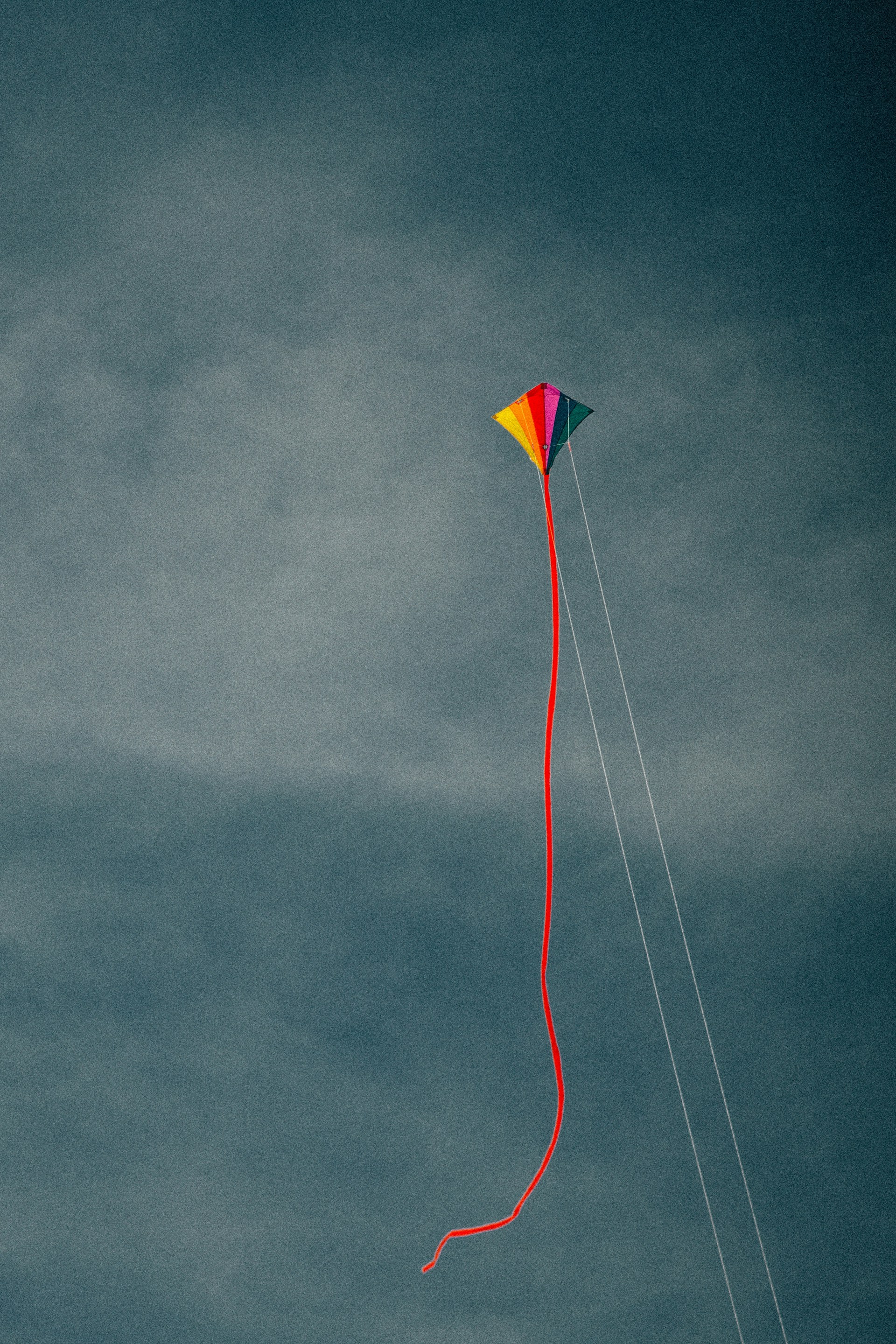 A colorful diamond-shaped kite with a long red tail flies high against a moody, dark grey overcast sky, held by two thin white control strings.