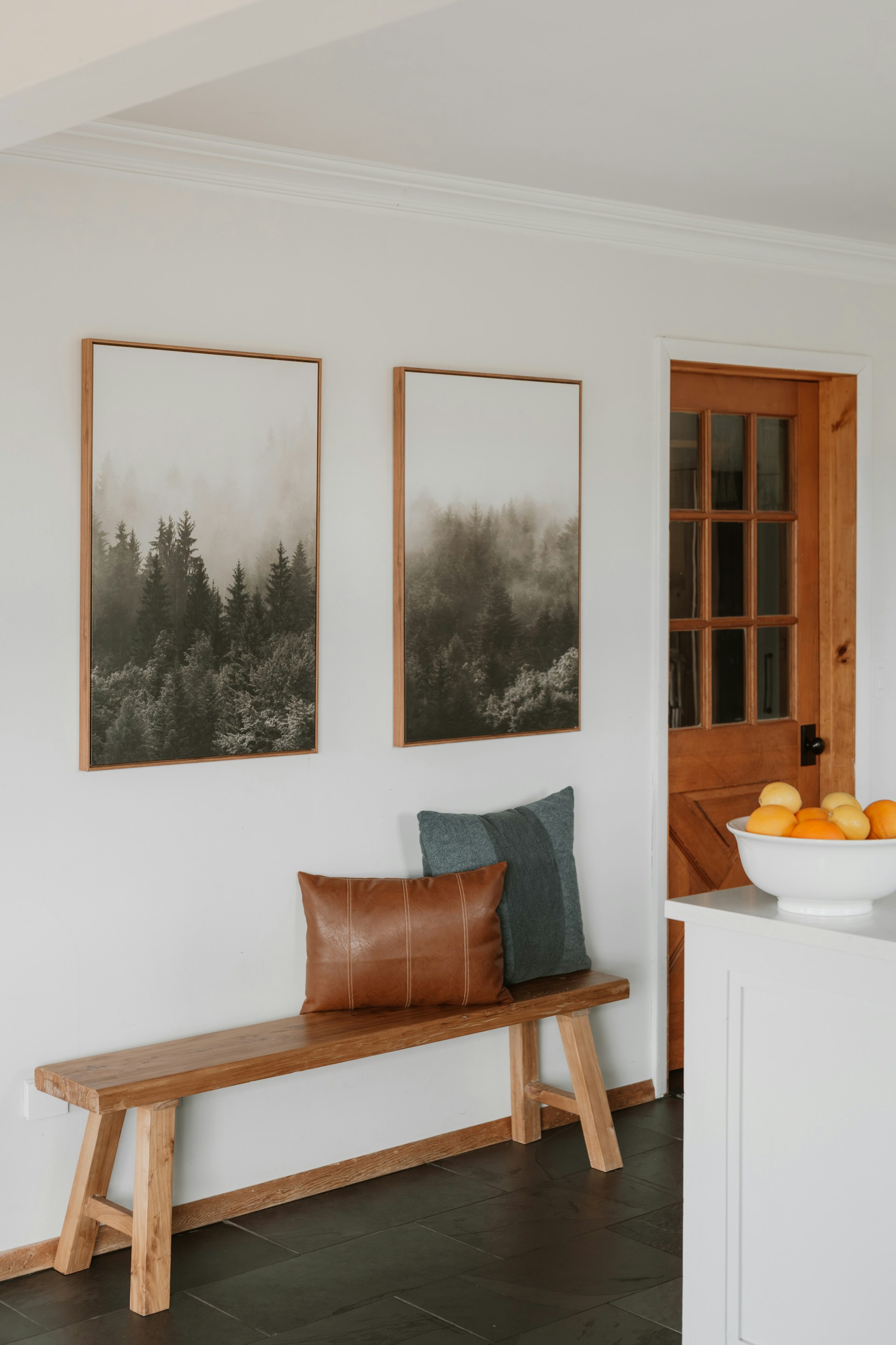 A wooden bench with a brown leather pillow and a blue cushion sits below two framed forest landscape prints on a white wall near a wooden door.