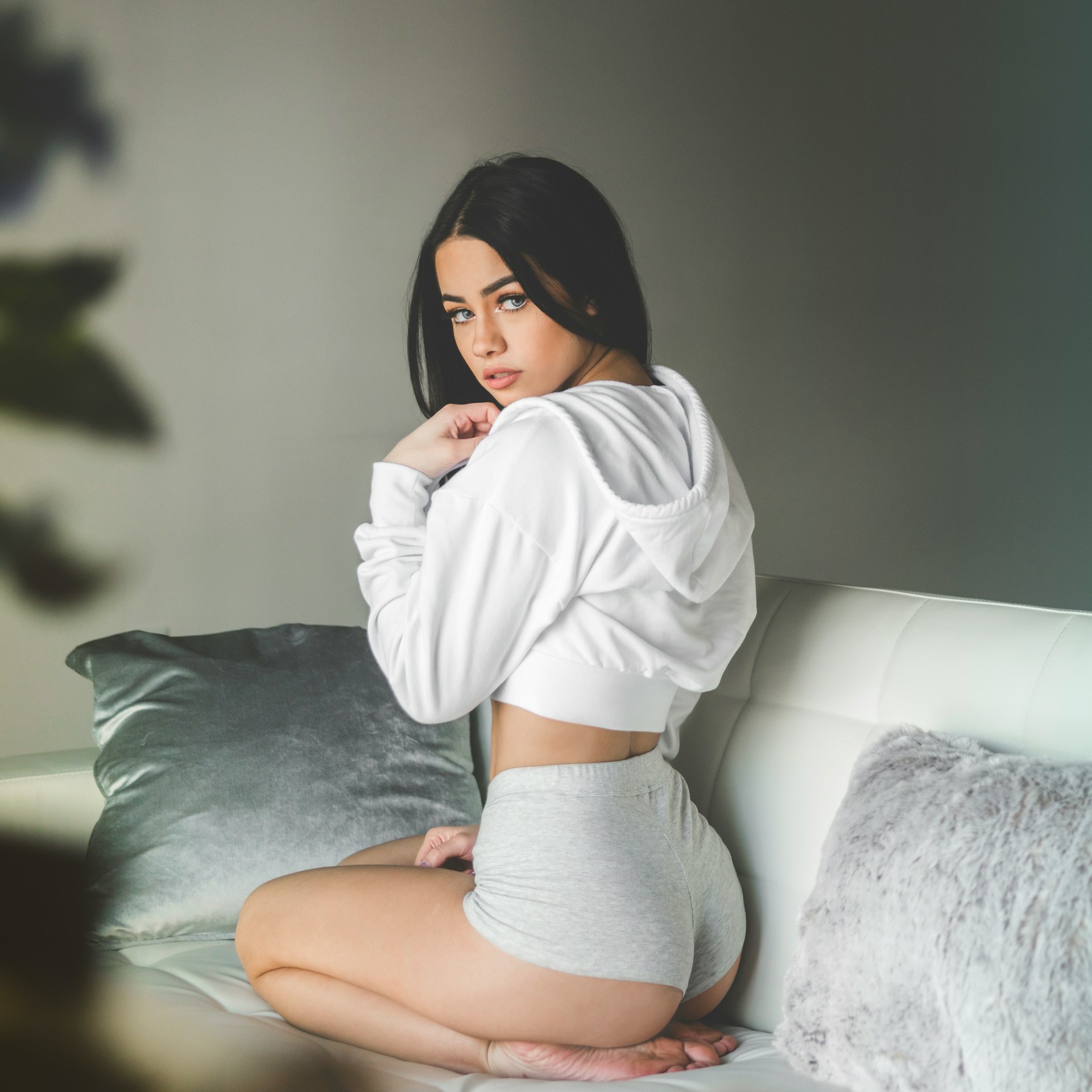 A young woman with dark hair wearing a white cropped hoodie and grey shorts kneels on a white sofa, looking back over her shoulder at the camera.