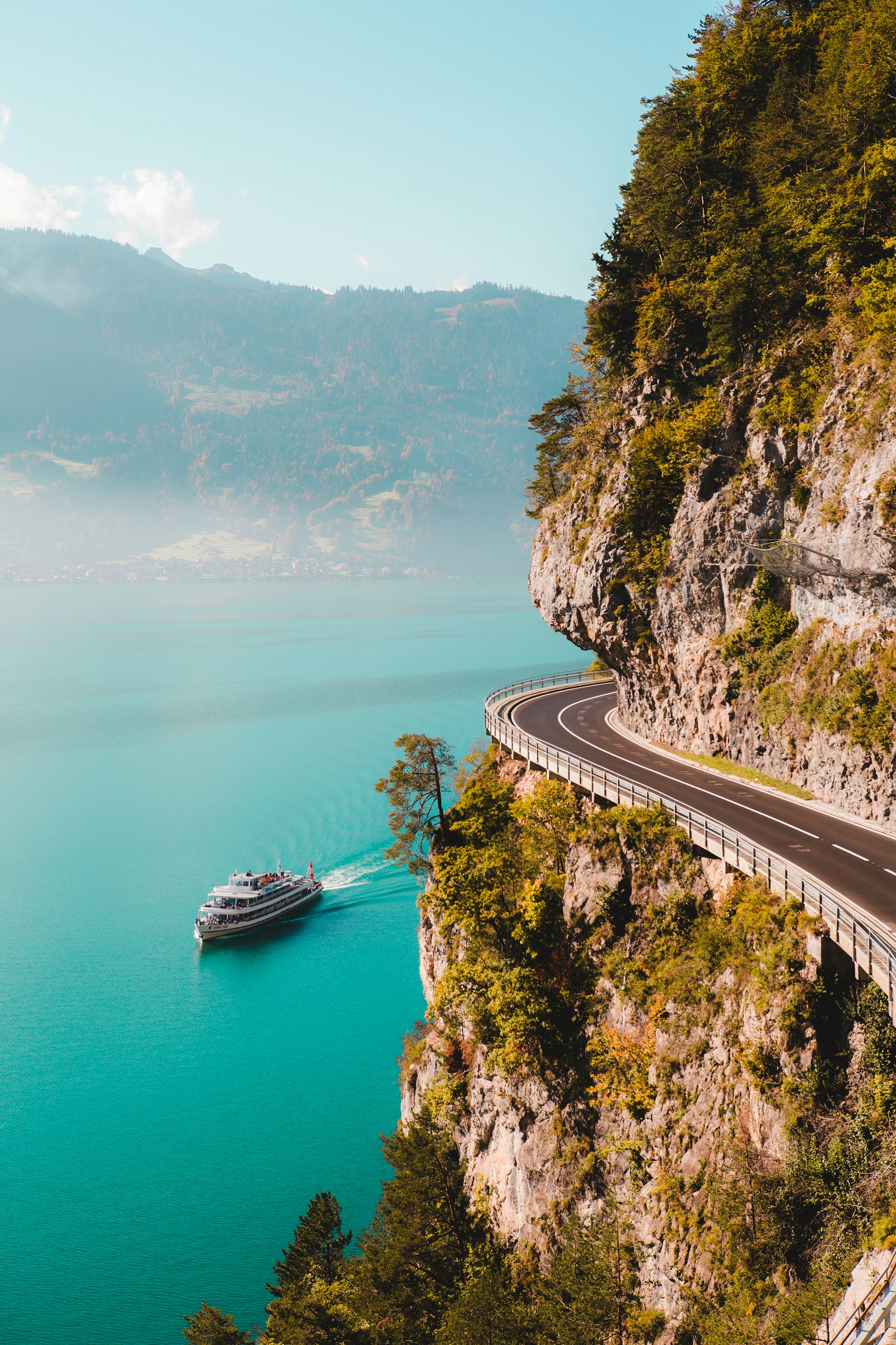 A scenic view of a passenger boat cruising on a bright turquoise lake next to a steep, rocky cliff with a winding road built along the mountainside.