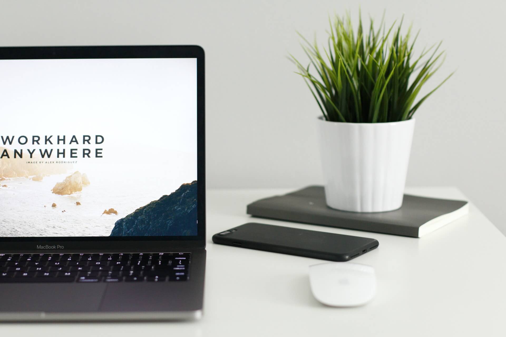 A MacBook Pro sits on a white desk next to a potted plant, a black smartphone, and a white computer mouse, with a coastal wallpaper on the screen.