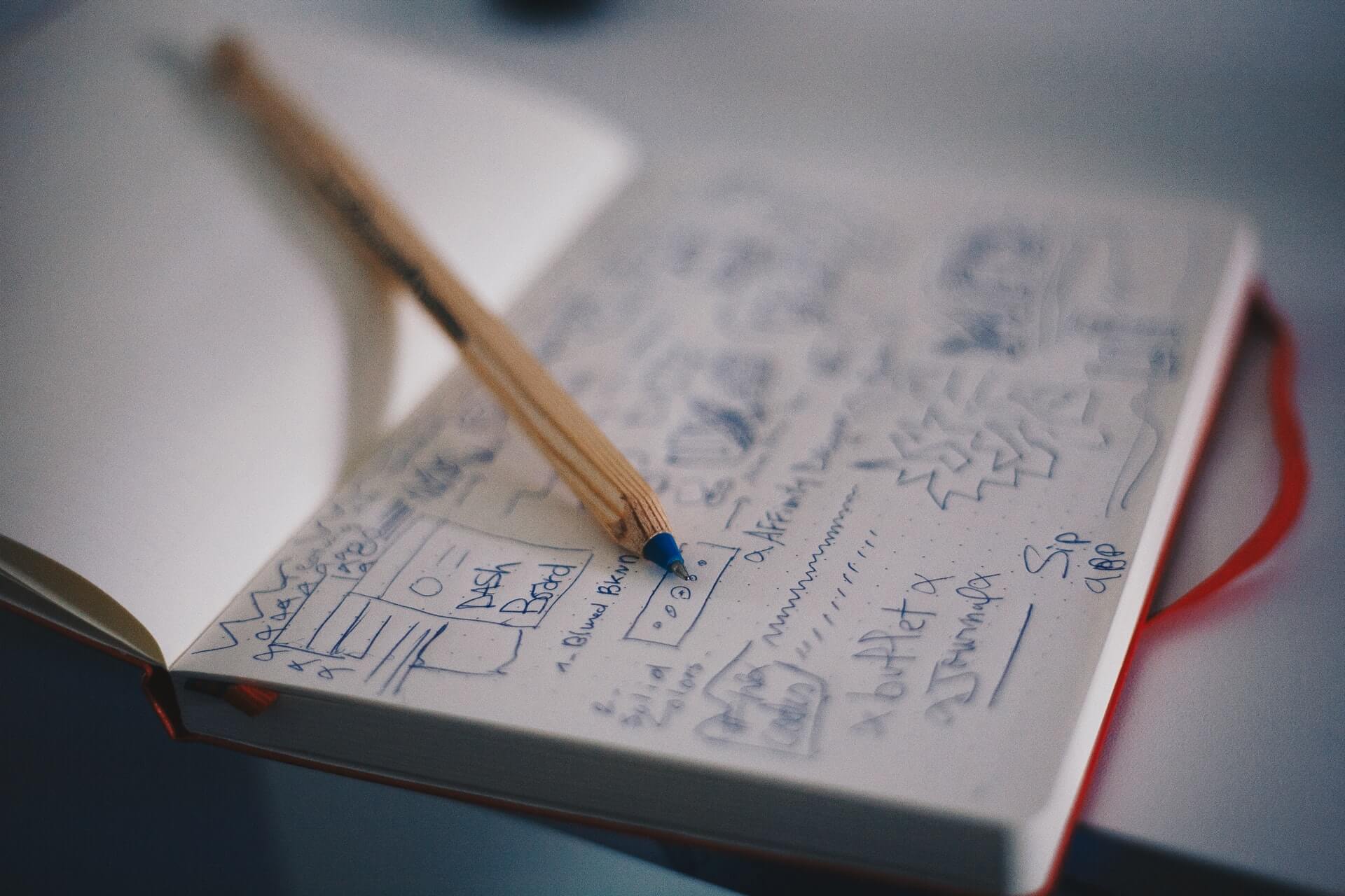 A wooden pencil rests on an open notebook filled with handwritten notes and rough sketches of a dashboard layout, captured in a soft-focus view.