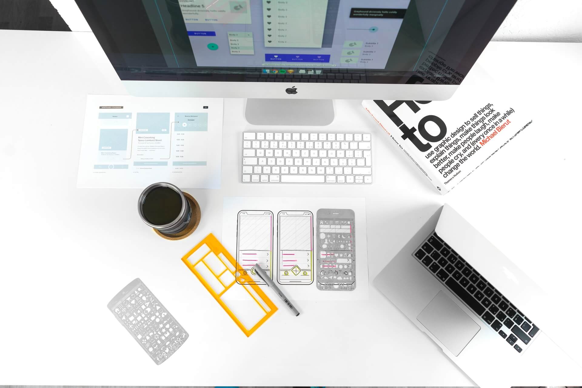 A high-angle view of a designer's white desk with a computer, laptop, sketches, a coffee mug, a design book, and various drafting stencils for UI.