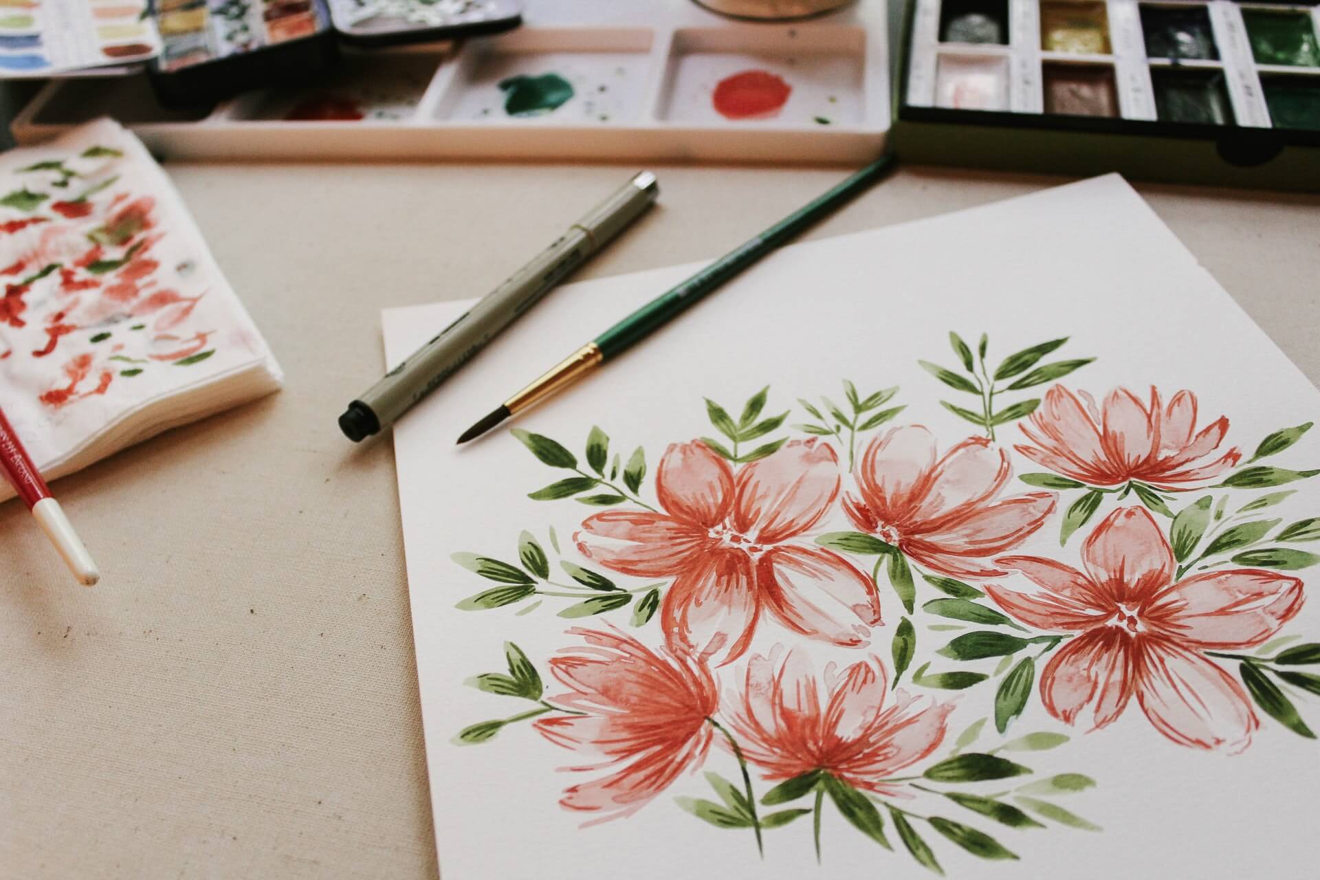 A watercolor painting of pink flowers and green leaves on white paper, with paintbrushes and a palette of watercolors arranged on a desk nearby.
